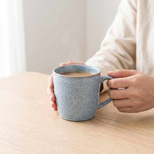 Ice Blue Nordic ceramic mug cradled in two hands on a pale oak table